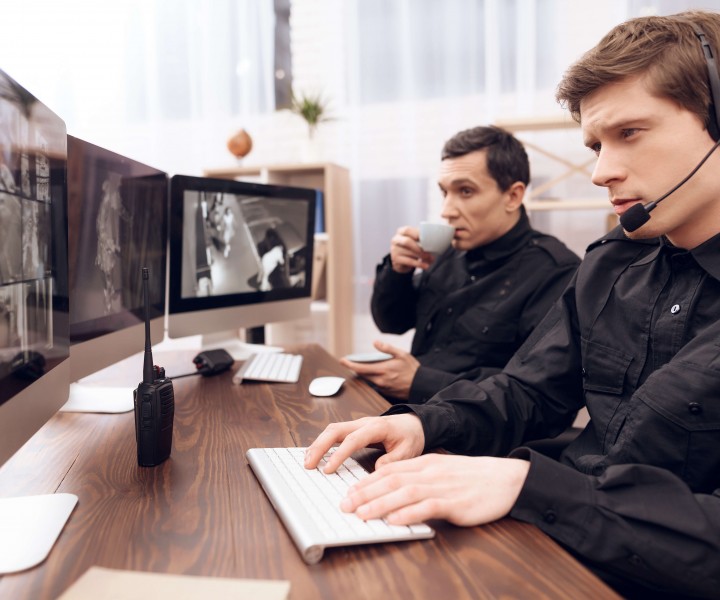 Two men work as guards. They sit in front of the monitors in the security room. The headphones are on their heads.